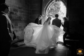 Back view of the bride’s dress billowing in the wind as she and the groom exit St Peter’s Church in Grandborough, moving from the dark interior to the bright outdoors.